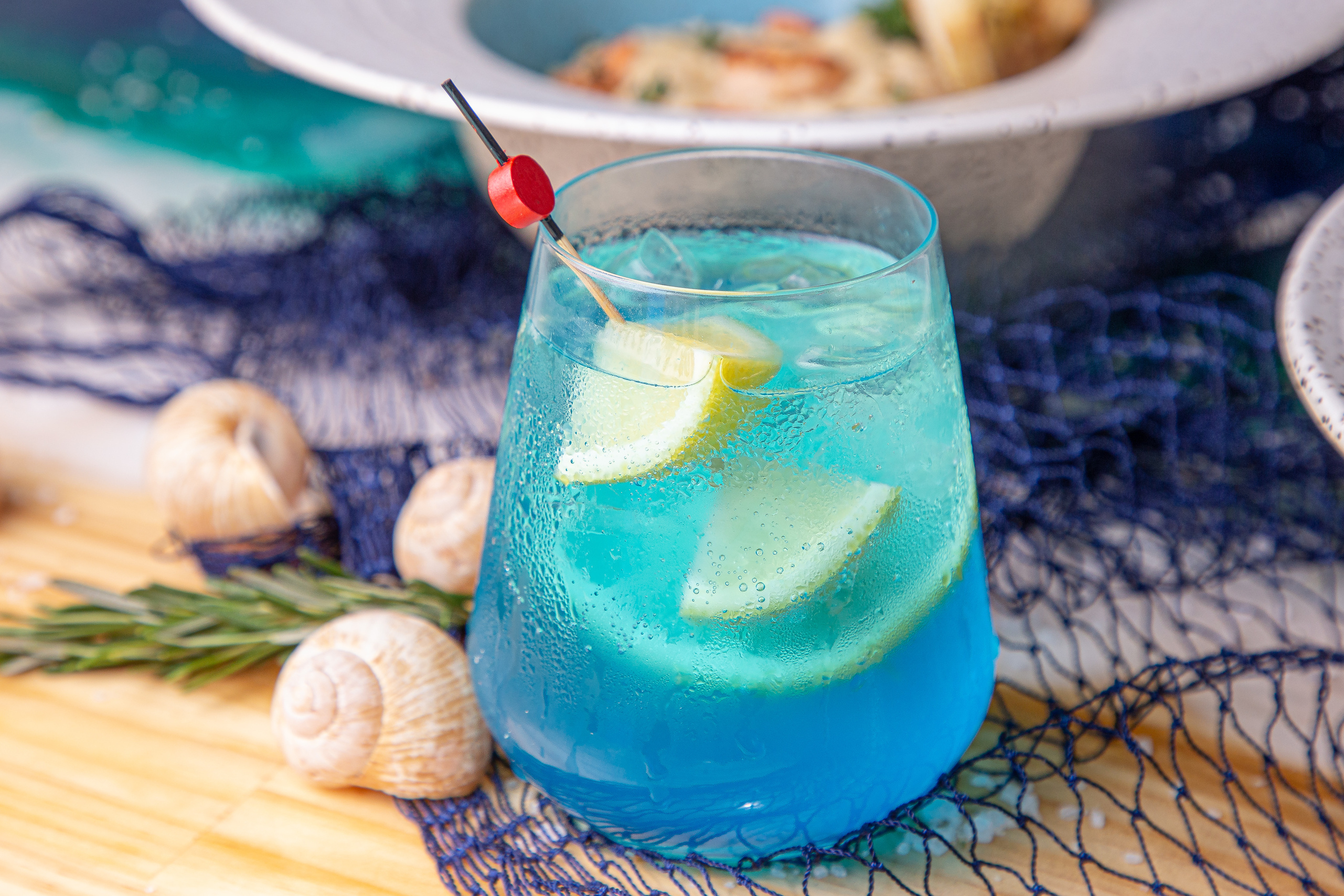 Drinque azul com limão e alecrim, em mesa com rede de pesca e conchas